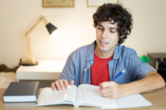 Young man studying and reading textbooks at his desk in his bedroom. Young man preparing for an exam.