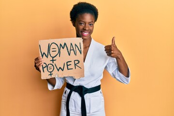 Young african american girl wearing karate kimono holding woman power banner smiling happy and positive, thumb up doing excellent and approval sign © Krakenimages.com