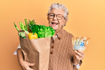 Senior grey-haired woman holding groceries and swiss franc banknotes winking looking at the camera...