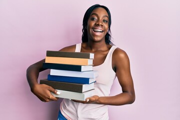 Young african american woman holding books celebrating crazy and amazed for success with open eyes screaming excited.