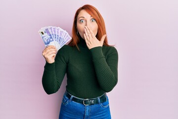 Beautiful redhead woman holding 20 swedish krona banknotes covering mouth with hand, shocked and afraid for mistake. surprised expression