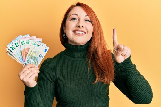 Beautiful redhead woman holding euro banknotes smiling with an idea or question pointing finger with happy face, number one