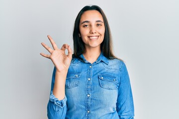 Young hispanic woman wearing casual denim jacket smiling positive doing ok sign with hand and...