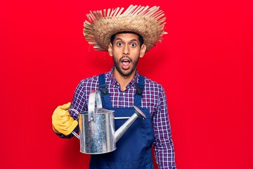 Young latin man wearing farmer hat and gloves holding watering can scared and amazed with open mouth for surprise, disbelief face © Krakenimages.com