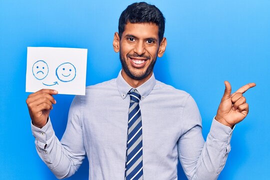 Young latin man holding sad to happy emotion paper smiling happy pointing with hand and finger to the side