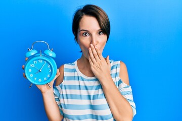 Young brunette woman with short hair holding alarm clock covering mouth with hand, shocked and afraid for mistake. surprised expression