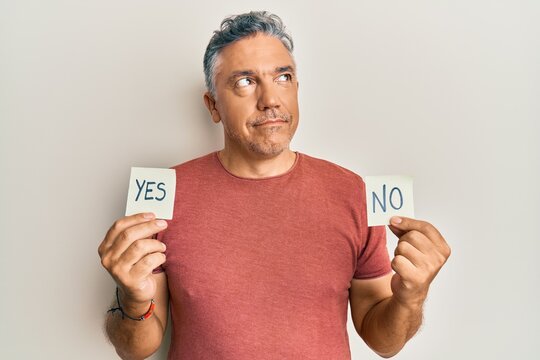 Handsome middle age mature man holding yes and no reminder smiling looking to the side and staring away thinking.
