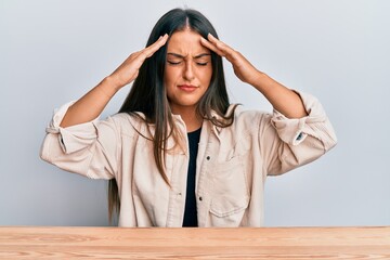Beautiful hispanic woman wearing casual clothes sitting on the table with hand on head, headache...