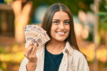 Young hispanic girl smiling happy holding mexican 500 pesos banknotes at the park. - obrazy, fototapety, plakaty