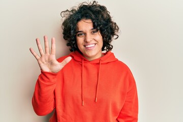 Young hispanic woman with curly hair wearing casual sweatshirt showing and pointing up with fingers number five while smiling confident and happy.