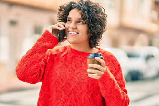 Young hispanic woman talking on the smartphone and drinking coffee at the city