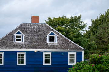 A vintage blue wooden building with white trim, double hung windows, dormers, a black shingled roof, and a brown brick chimney. The old home has multiple stories with a green tree and grey skies.