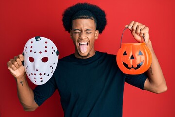African american man with afro hair wearing hockey mask and halloween pumpking sticking tongue out happy with funny expression.