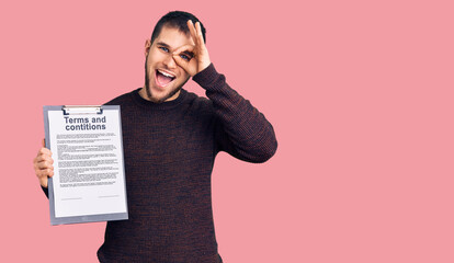 Young handsome man holding clipboard with terms and conditions document smiling happy doing ok sign with hand on eye looking through fingers