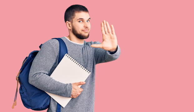 Young handsome man wearing student backpack holding notebook with open hand doing stop sign with serious and confident expression, defense gesture
