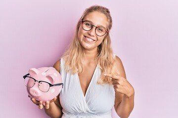 Young blonde girl holding piggy bank with glasses pointing finger to one self smiling happy and...