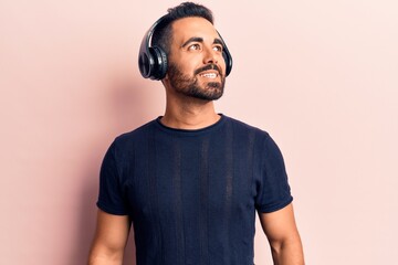 Young hispanic man listening to music using headphones looking away to side with smile on face,...