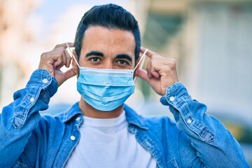 Young hispanic man putting on medical mask standing at the city.