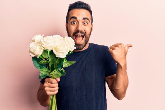 Young hispanic man holding flowers pointing thumb up to the side smiling happy with open mouth