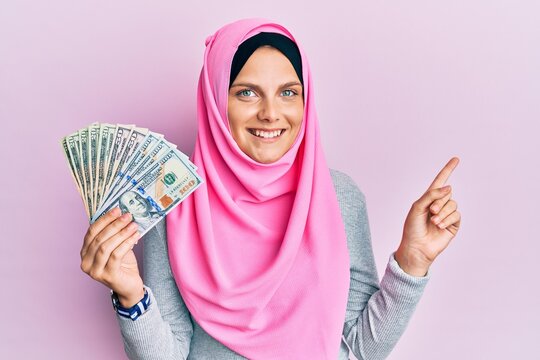 Young caucasian woman wearing islamic hijab holding dollars banknotes smiling happy pointing with hand and finger to the side
