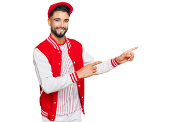 Young man with beard wearing baseball uniform smiling and looking at the camera pointing with two hands and fingers to the side.