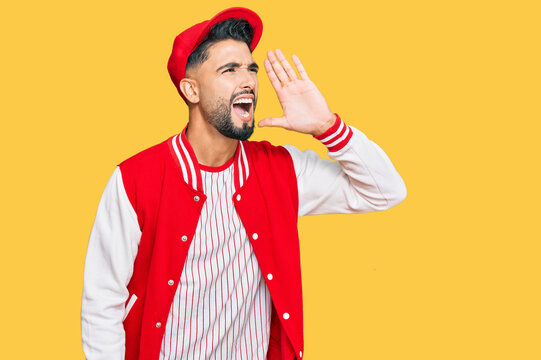Young man with beard wearing baseball uniform shouting and screaming loud to side with hand on mouth. communication concept.