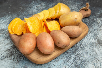 Top view of crispy chips and uncooked potatoes on wooden cutting board on gray background
