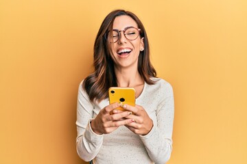 Young brunette woman using smartphone over yellow background smiling and laughing hard out loud...