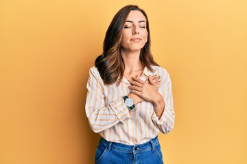 Young brunette woman wearing casual clothes over yellow background smiling with hands on chest, eyes closed with grateful gesture on face. health concept.