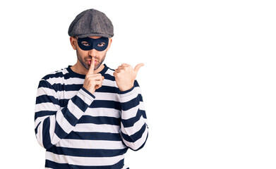 Young handsome man wearing burglar mask asking to be quiet with finger on lips pointing with hand to the side. silence and secret concept.