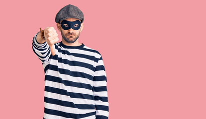 Young handsome man wearing burglar mask looking unhappy and angry showing rejection and negative with thumbs down gesture. bad expression.