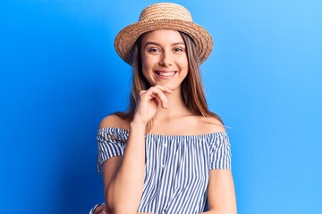 Young beautiful girl wearing summer hat and striped t-shirt smiling looking confident at the camera with crossed arms and hand on chin. thinking positive.