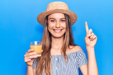 Young beautiful girl wearing summer hat drinking glass of orange juice smiling with an idea or question pointing finger with happy face, number one