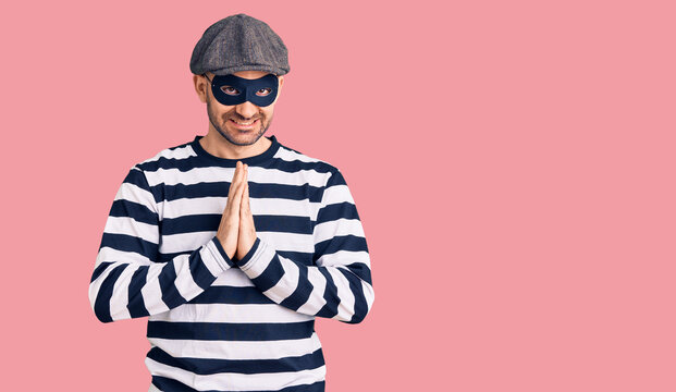 Young handsome man wearing burglar mask praying with hands together asking for forgiveness smiling confident.