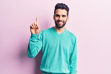 Fototapeta premium Young handsome man with beard wearing casual sweater smiling with an idea or question pointing finger up with happy face, number one