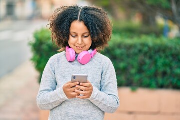 Adorable hispanic child girl listening to music using smartphone and headphones at the park.