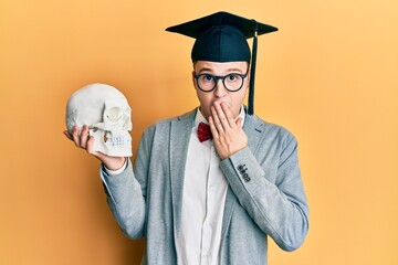 Young caucasian nerd man wearing glasses and graduation cap holding skull covering mouth with hand,...
