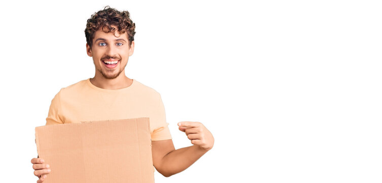 Young handsome man with curly hair holding blank cardboard empty banner smiling happy pointing with hand and finger