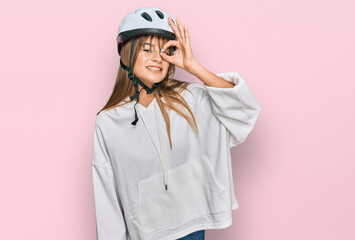 Teenager caucasian girl wearing bike helmet doing ok gesture with hand smiling, eye looking through fingers with happy face.