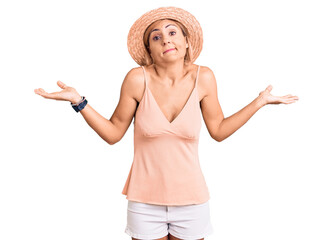 Young blonde woman wearing summer hat clueless and confused expression with arms and hands raised....