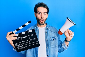 Young hispanic man holding video film clapboard and megaphone afraid and shocked with surprise and...