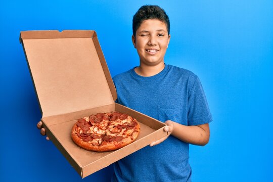 Teenager hispanic boy eating tasty pepperoni pizza winking looking at the camera with sexy expression, cheerful and happy face.