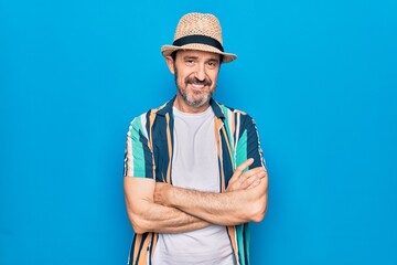 Middle age handsome tourist man on vacation wearing shirt and hat over blue background happy face smiling with crossed arms looking at the camera. Positive person.