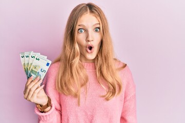 Beautiful young caucasian girl holding bunch of 5 euro banknotes scared and amazed with open mouth...