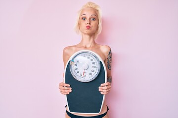 Young blonde woman with tattoo standing shirtless holding weighing machine puffing cheeks with funny face. mouth inflated with air, catching air.