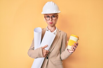 Young blonde woman with tattoo holding paper blueprints drinking take away cup of coffee depressed...