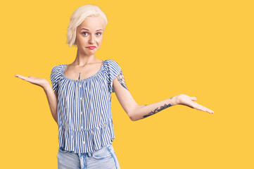 Young blonde woman with tattoo wearing casual clothes clueless and confused expression with arms and hands raised. doubt concept.