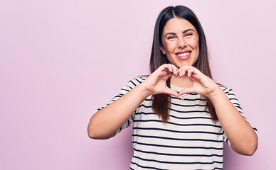 Young beautiful brunette woman wearing casual striped t-shirt over isolated pink background smiling in love doing heart symbol shape with hands. Romantic concept.