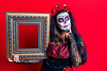 Young woman wearing day of the dead costume holding empty frame serious face thinking about question with hand on chin, thoughtful about confusing idea