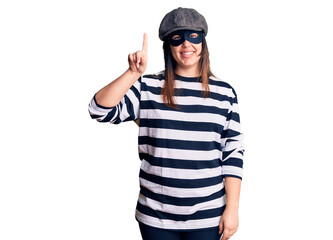 Young beautiful brunette woman wearing burglar mask showing and pointing up with finger number one while smiling confident and happy.